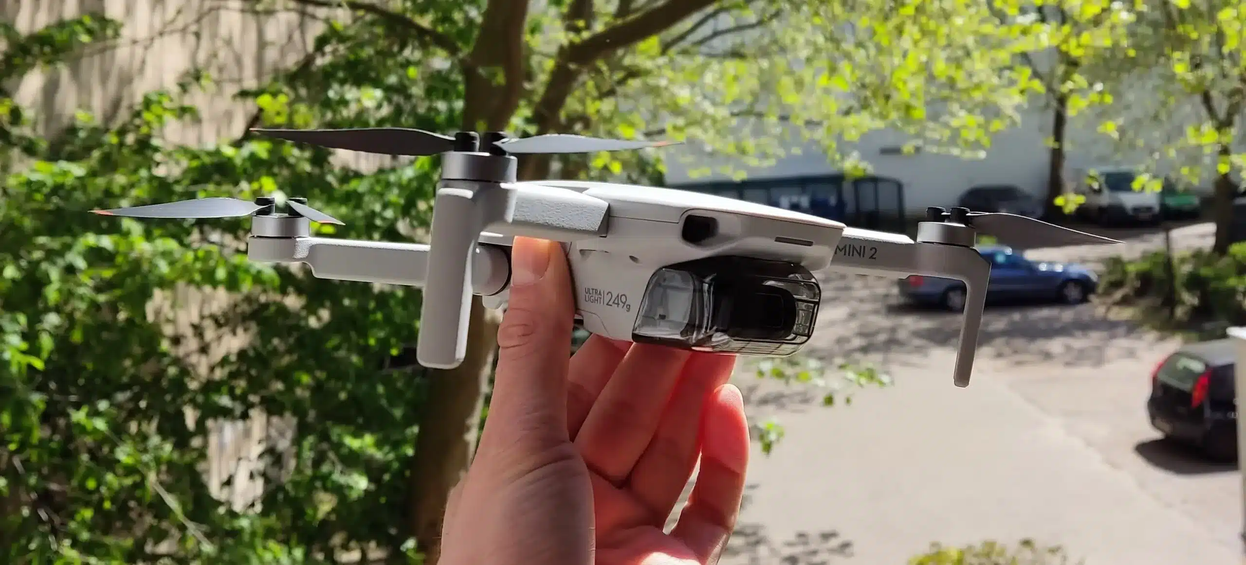 Drohne DJI Mini 2 wird in der Hand gehalten im Hintergrund Bäume und ein Parkplatz